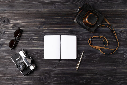 Vintage camera, sunglasses, and an open notebook with a pen are arranged on a rustic wooden surface, creating a nostalgic atmosphere for photography enthusiasts and creatives