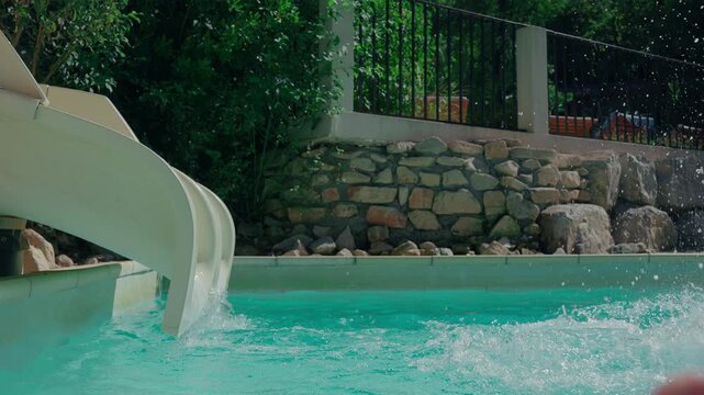 An 8-year-old girl splashes into a swimming pool from a waterslide wearing goggles. The scene is captured in slow motion