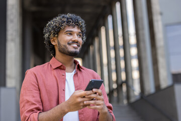 Close-up photo of a young smiling Indian man standing on the street, holding a phone and looking ahead