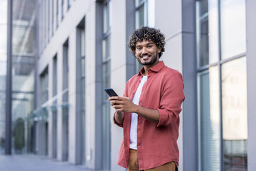 Portrait of a young Muslim man in a red shirt standing near a building on the street, holding a phone in his hand and looking at the camera