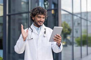 A young Indian doctor is standing on the street near the hospital, talking on a video call with a patient on a tablet, greeting and waving at the camera