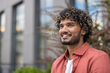 Close-up photo of a young Indian man in a red shirt sitting outside and looking to the side with a smile