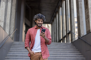 A young Indian man is walking down the stairs of an office building, holding his left hand in his pocket and looking at his mobile phone screen