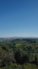 Obraz premium Scenic panoramic view from San Gimignano, Tuscany, Italy. Rolling green hills, vineyards, and cypress trees under a clear blue sky. Iconic Italian countryside landscape, travel, and nature background.