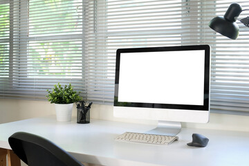 A white computer monitor mockup is placed on a table next to a window in an office.