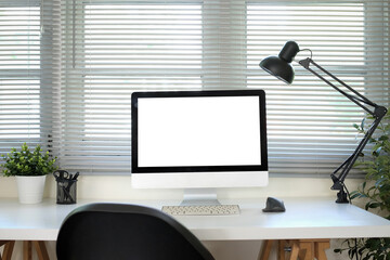 A white computer monitor mockup is placed on a table next to a window in an office.