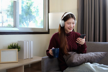 A young woman sits playing on her phone and wearing headphones in the living room of her house.