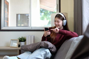 A young woman sits playing on her phone and wearing headphones in the living room of her house.