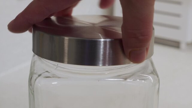 A hand firmly places a brushed metal lid onto a clear glass storage jar, sealing its contents. This highlights an act of organizing and preserving food or items in a kitchen.