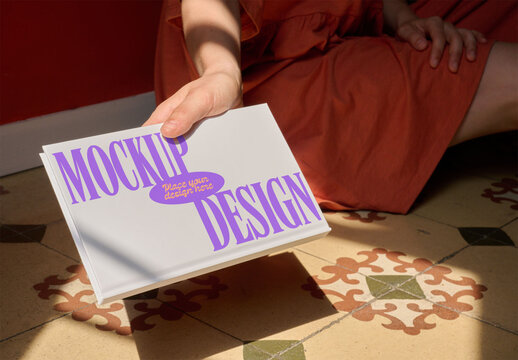 A female hand presents a blank notebook while seated on a colorful tiled floor. PSD template mockup