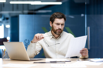 A frustrated young man sits at a desk in the office, holding glasses in his hand and looking incomprehensibly at and studying documents