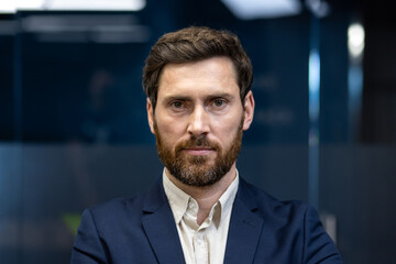 Close-up portrait of a young businessman in a suit standing in an office and looking seriously at the camera