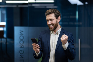 Close-up photo of a young happy man standing in a company office, looking at the phone screen and showing a victory sign with his hand
