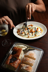 Young woman having dinner with appetizer of anchovies, cream sauce, and croutons on a white plate, with a martini glass holding a green olive and a bread basket tray also on the dark wooden table