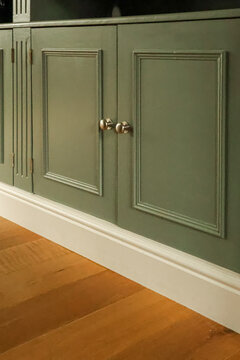 Green wooden cabinet with panel doors and brass knobs, white baseboard accenting hardwood flooring in a home interior