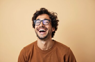 Obraz premium Young man with curly hair and glasses laughs heartily against a plain beige background. He wears a brown sweater and shows a wide, joyful smile, conveying happiness and confidence.