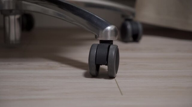 A close-up view captures a dark gray caster wheel, attached to a shiny silver metal chair base, resting firmly on a light-colored wooden floor, highlighting the mobility and stability