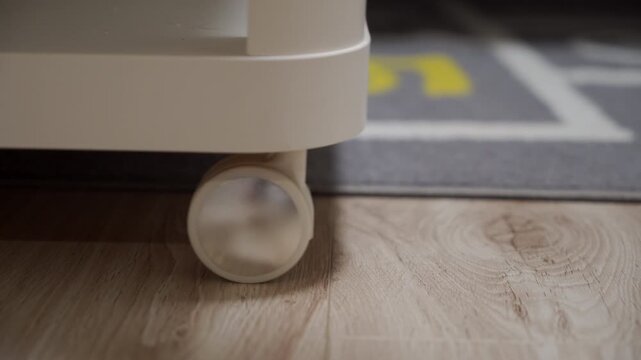 A close-up view highlights a white caster wheel, part of a piece of furniture on a light wooden floor, with a blurred gray rug and a hint of yellow color visible in the background