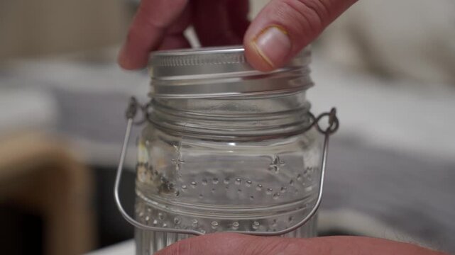 Human hands are seen interacting with a clear glass jar featuring a silver metallic lid and a wire bail handle, suggesting an action of opening or closing the container.
