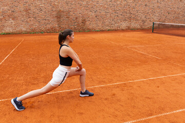 Obraz premium Woman stretching legs on clay tennis court. Concept of sports training, flexibility, preparation and physical wellness through outdoor athletic activity.