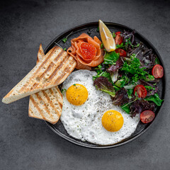 Breakfast with fried eggs, salmon, and salad top view. Top view of breakfast with fried eggs, salmon, red caviar, green salad, and toasted bread on a dark plate.
