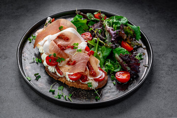 Bruschetta with prosciutto and salad greens top view. Top view of bruschetta with prosciutto, cherry tomatoes, cream cheese, and salad greens served on dark plate.