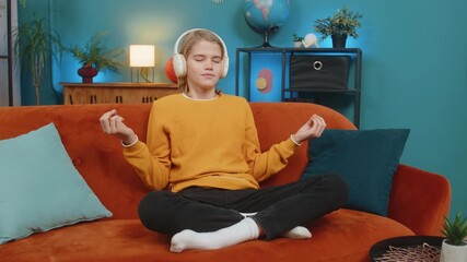 Caucasian teenager boy meditates in lotus pose on comfortable sofa at home. Wearing headphones listening music, child relaxes unwinds peacefully after long school day, finding inner peace calmness