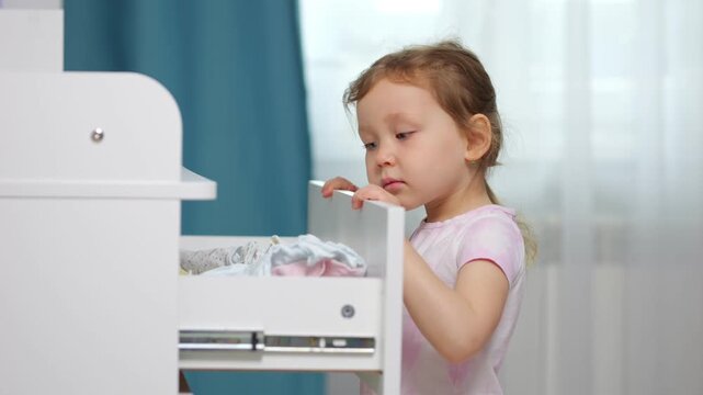 Small girl pulls open white dresser drawer with baby clothes