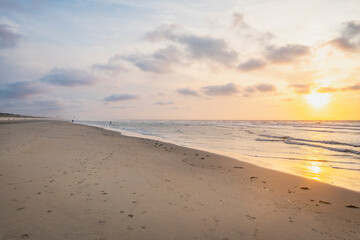 Sunset over stormy North Sea beach with reed grass bushes, orange-red sky over the dark blue ocean, panorama with sandy beach and blowing grass in the wind, storm at sunset over wavy water