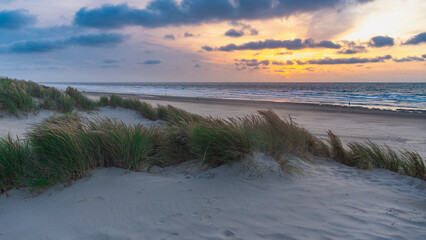 Sunset over stormy North Sea beach with reed grass bushes, orange-red sky over the dark blue ocean, panorama with sandy beach and blowing grass in the wind, storm at sunset over wavy water