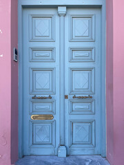 Vintage blue wooden door with brass handles and geometric panels framed by pink walls. Traditional Mediterranean entrance, architectural detail from Greece, pastel color contrast.