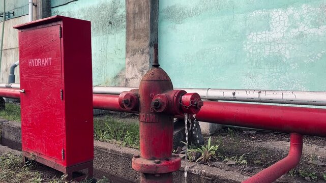 An old, red fire hydrant leaks water near a matching cabinet in an industrial setting. The hydrant shows signs of rust and wear, indicating a need for maintenance or repair.