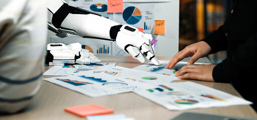 A robotic arm collaborates with a human in an office setting, analyzing business metrics and data presented through colorful graphs and reports to enhance productivity.Noogenesis