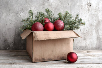 Christmas decorations in a cardboard box on a rustic wooden table. Red holiday ornaments and fir branches against a gray background.
