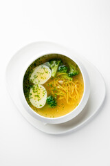 Clear vegetable soup with broccoli, potato slices, noodles, and herbs in a white bowl on a plate. Minimalist presentation, healthy homemade meal, top view.