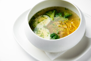 Clear vegetable soup with broccoli, potato slices, noodles, and herbs in a white bowl on a plate. Minimalist presentation, healthy homemade meal, top view.