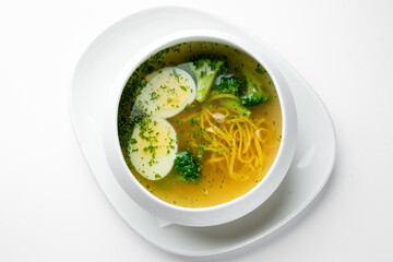 Clear vegetable soup with broccoli, potato slices, noodles, and herbs in a white bowl on a plate. Minimalist presentation, healthy homemade meal, top view.