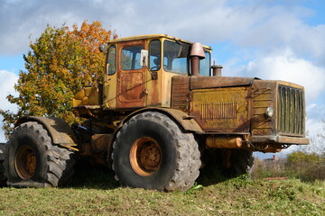 Fototapeta premium Rusted Vintage Soviet Articulated Tractor on Grassy Hillside with Autumn Tree