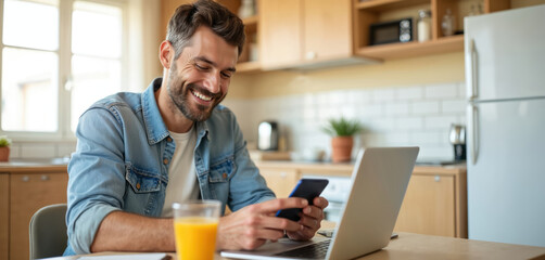 Man smiles while using smartphone near laptop with orange juice in kitchen. He is happy about online banking or ecommerce transaction with credit card at home.
