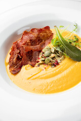 Gourmet pumpkin soup with crispy bacon, seeds, and fresh herbs served in a white bowl. Autumn-themed dish with elegant plating, ideal for restaurant or seasonal food concepts.