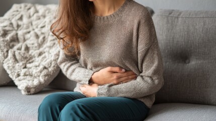 Obraz premium Woman sitting on a sofa experiencing stomach pain while wearing a cozy sweater