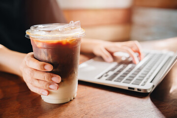 Coffee concept. Close-up of Hand Holding Iced Coffee Cup While Using Laptop at Cozy Cafe Table with Natural Light and Warm Atmosphere