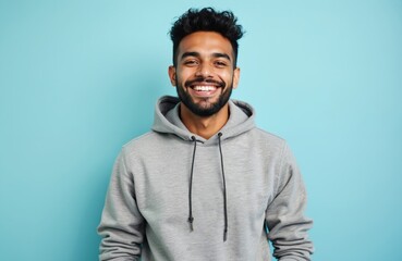 Obraz premium Young man with beard, dark curly hair, smiles broadly. Wears casual grey hoodie on light blue background. Person looks confident, cheerful, positive. Young adult male feels joy, good mood. Guy