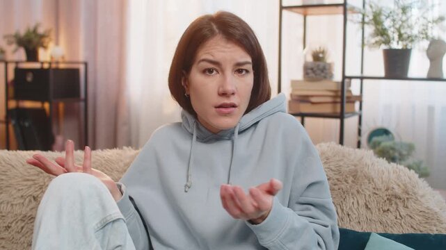 Confused Caucasian woman raising eyebrows feeling embarrassed about ambiguous question, having doubts, no idea, being clueless and uncertain. Young adult girl sitting on sofa in living room at home