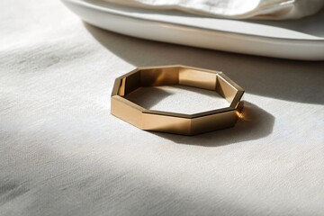 A stylish, gold octagonal bracelet rests on a linen tablecloth, illuminated by sunlight.