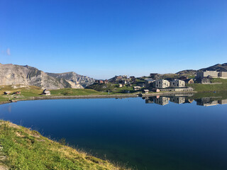 Klares Bergpanorama am Lac de Tignes in den franz&ouml;sischen Alpen, mit spiegelglatter Wasseroberfl&auml;che, modernen Geb&auml;uden und blauen Himmel &ndash; friedliche alpine Morgenstimmung.