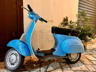 Hellblauer Vintage-Roller steht vor einer alten Hauswand auf Kopfsteinpflaster in mediterraner Altstadt bei Abendlicht. Nostalgisches Retro-Feeling mit s&uuml;dl&auml;ndischem Flair.