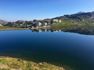 Klares Bergpanorama mit spiegelglattem See auf einem sonnigen Hochplateau, umgeben von gr&uuml;nen Wiesen und alpinen Geb&auml;uden unter strahlend blauem Himmel.