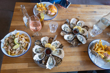 Seafood meal on a wooden table. Seen from above. There are rock oysters, fries with a crab dressing and lobster rolls with chips. A glass of rose is on the table and a man is holding a smart phone.