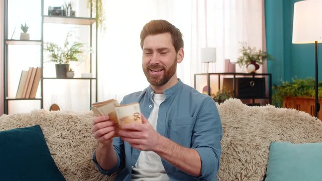 Young man at home holding a stack of euro cash banknotes, counting money with excitement. Caucasian guy on sofa smiles, feeling happy about a big paycheck lottery win or unexpected financial success